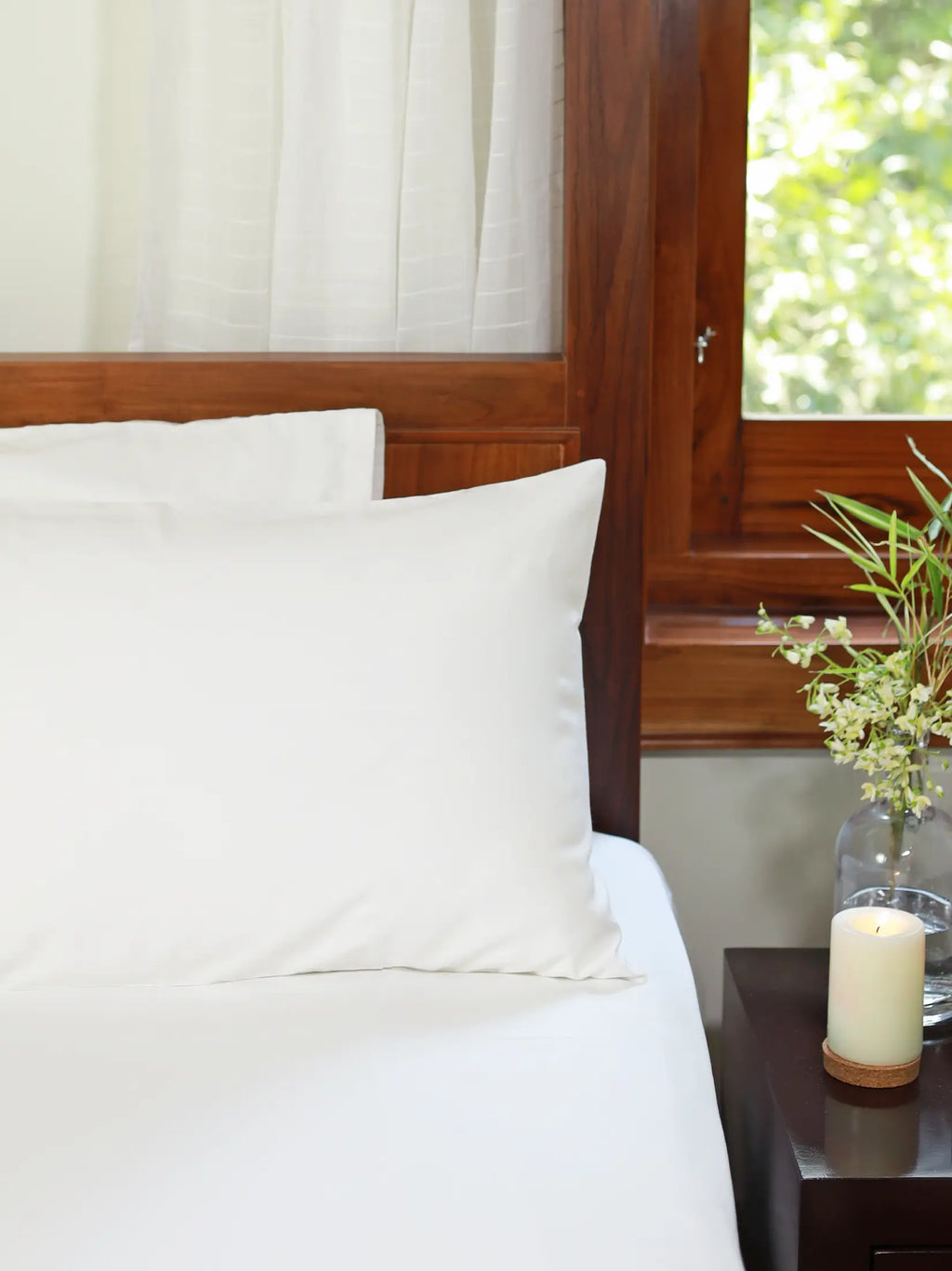 Plain white bed sheet and pillowcases on a bed beside a wooden nightstand
