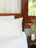 Plain white bed sheet and pillowcases on a bed beside a wooden nightstand