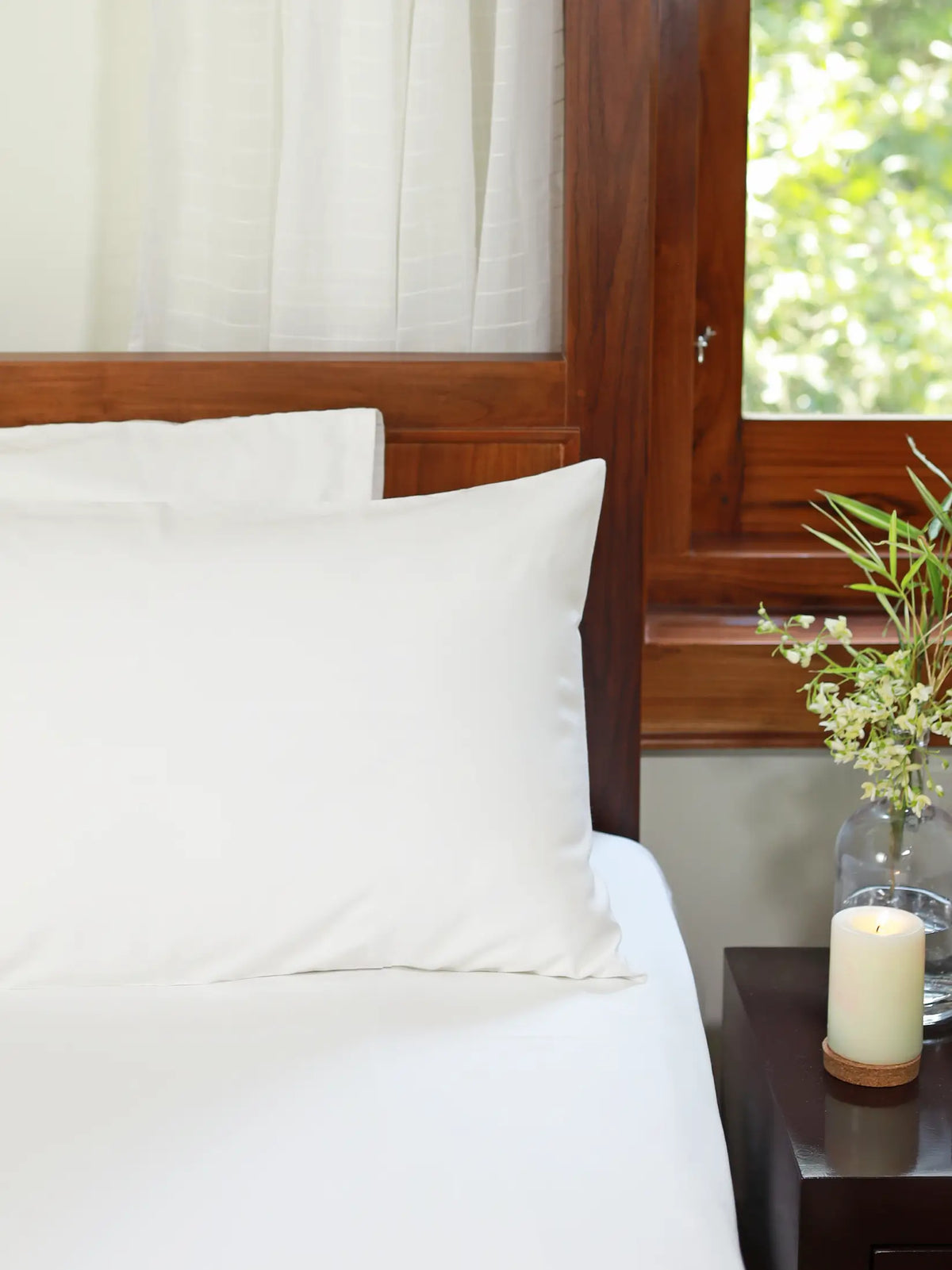 Plain white bed sheet and pillowcases on a bed beside a wooden nightstand
