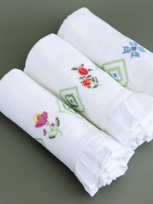 Three folded white Kerala bath towels with pink, red and blue embroidered motifs and ruffled edges