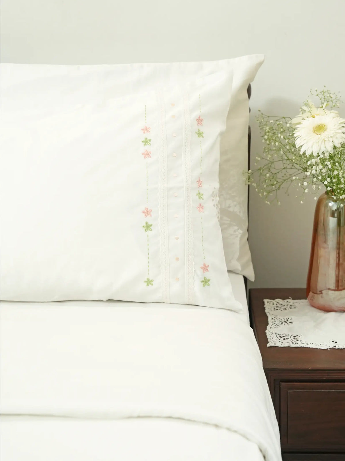 White embroidered pillowcase on a bed beside a vase of white flowers