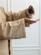 Woman applying lotion while wearing a beige waffle bath robe with embroidered cuffs