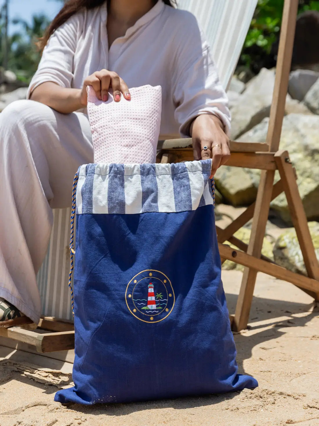 Woman placing a folded pink towel into a navy blue lighthouse-embroidered laundry bag on a beach