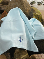 Pale blue waffle bath towel with navy anchor embroidery draped over seaside rocks
