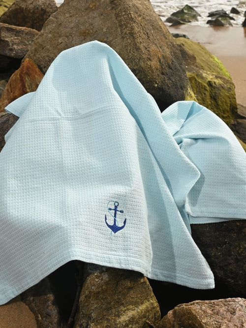 Pale blue waffle bath towel with navy anchor embroidery draped over seaside rocks