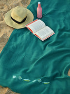 Teal green embroidered honeycomb towel on the beach with a straw hat, book and bottle