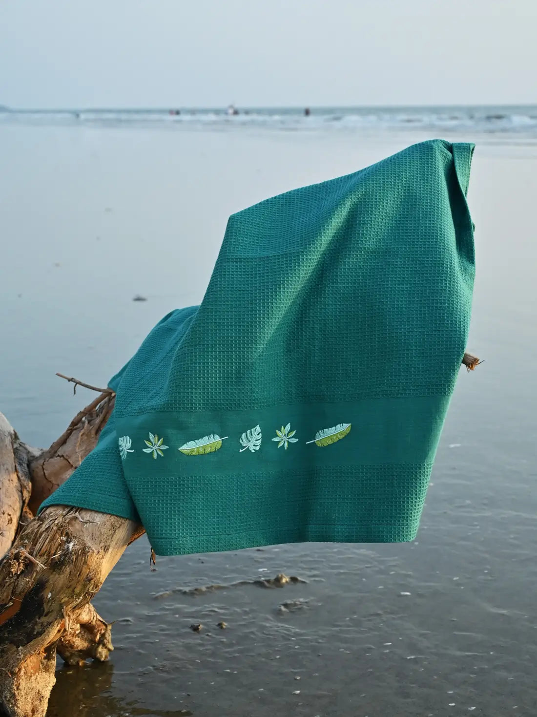 Teal green honeycomb towel with tropical leaf embroidery draped over driftwood at the beach