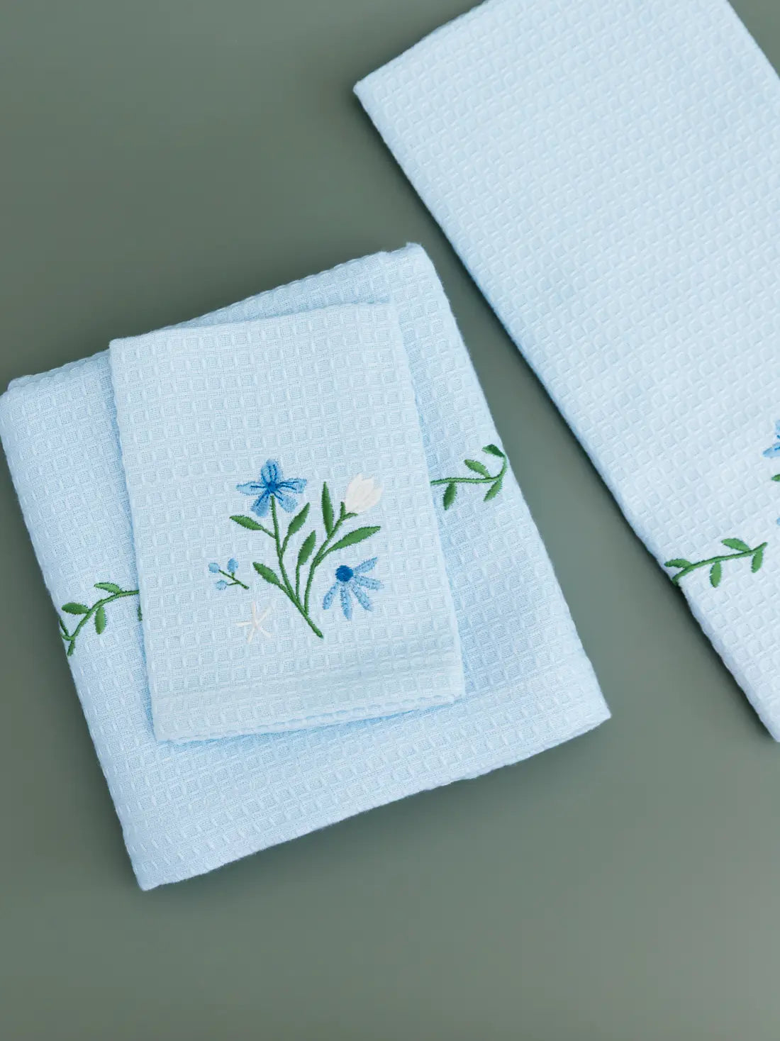 Three folded pale blue waffle towels with blue flowers and green vine embroidery
