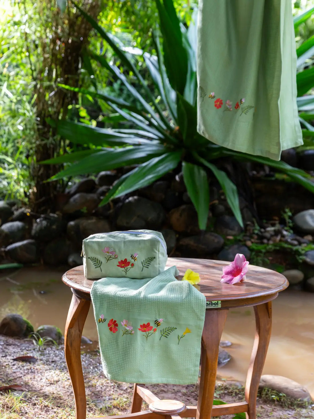 Olive green floral embroidered towel and matching zip pouch on a wooden stool by a stream