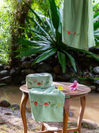 Olive green floral embroidered towel and matching zip pouch on a wooden stool by a stream