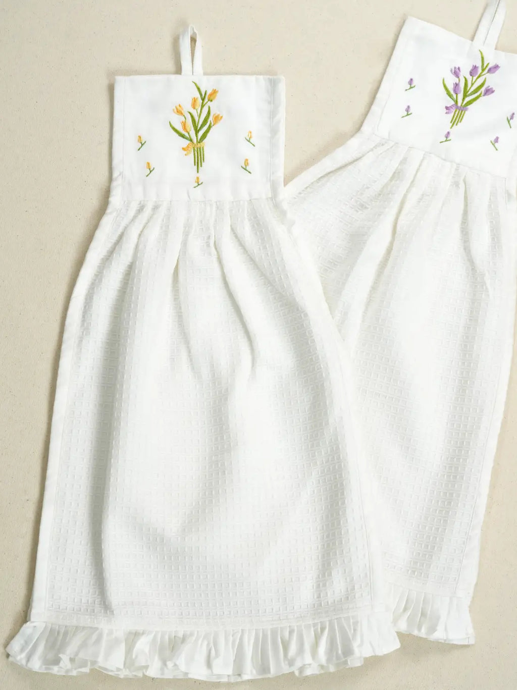 Two white hanging waffle hand towels with yellow and purple tulip embroidery and ruffled hems
