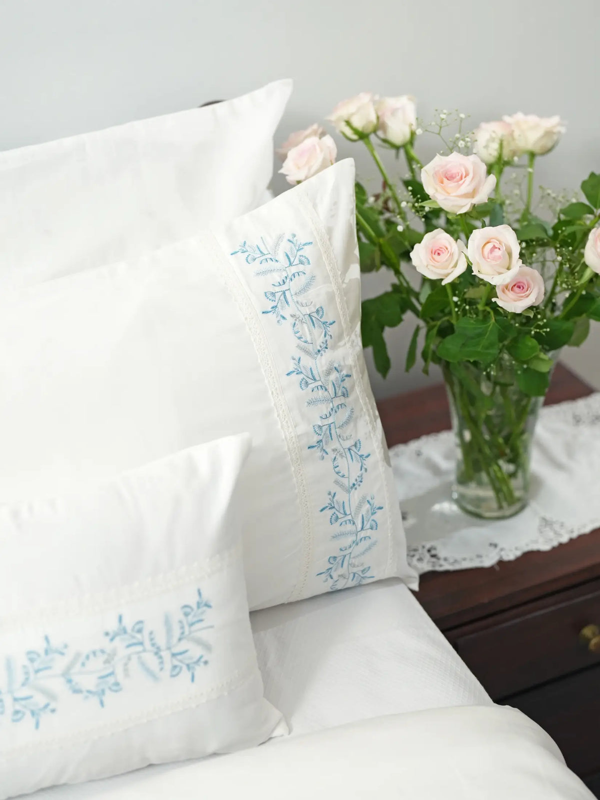 Close-up of a white pillowcase with blue vine embroidery beside pale pink roses